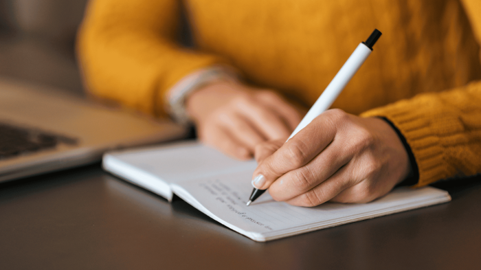 Person in a mustard sweater writing notes with a pen in a notebook on a table near a laptop.
